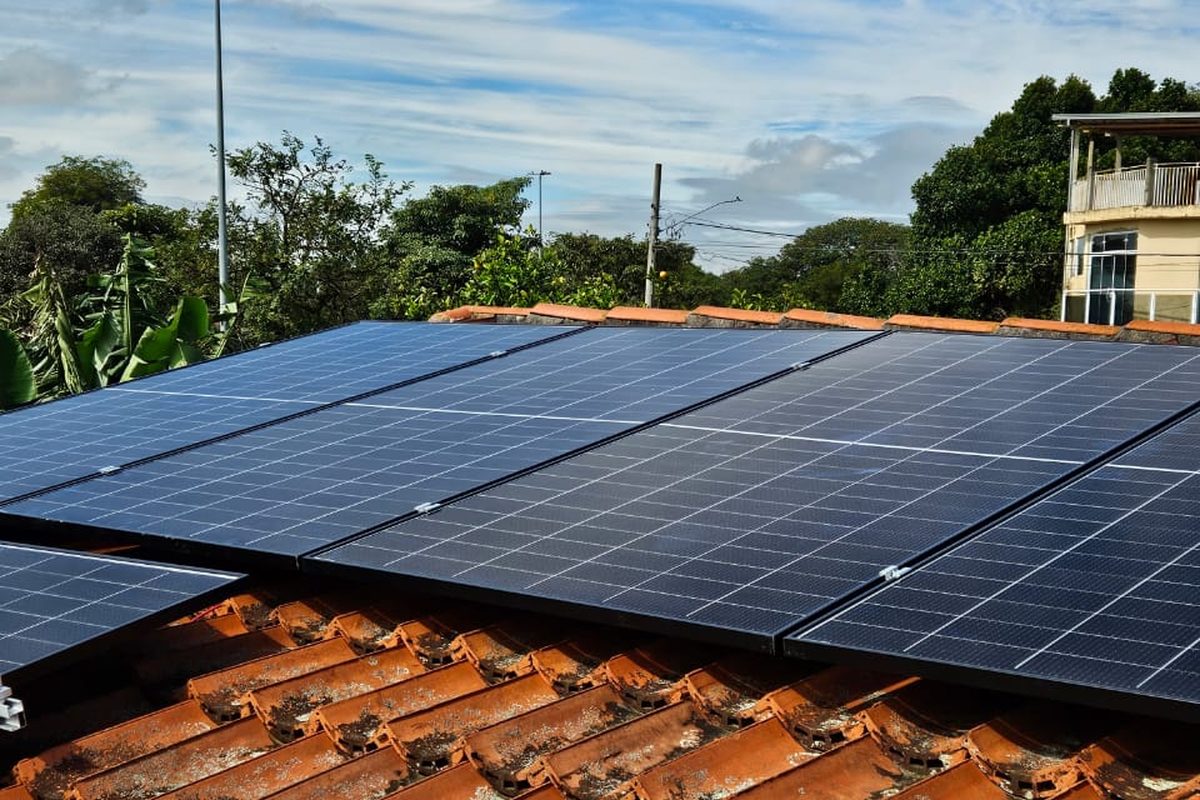 Vista ampla do telhado residencial com duas fileiras de módulos solares e
                  vegetação ao redor