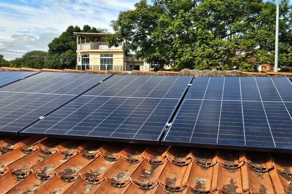 Fileira de módulos solares sobre telhado de telha cerâmica, com casa de dois
                  pavimentos ao fundo